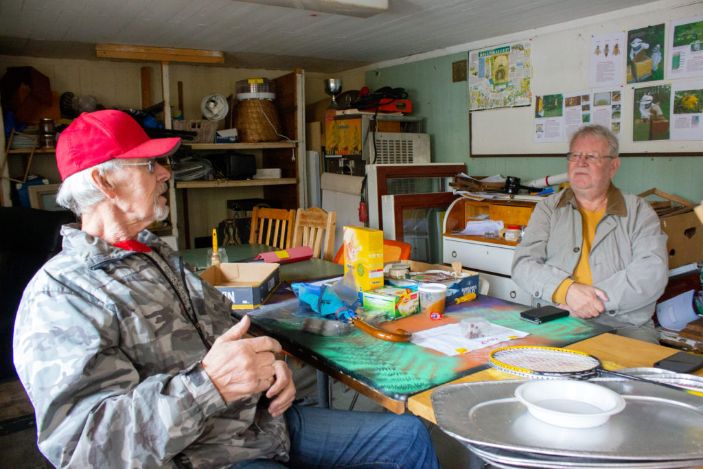Dennis och Lasse sitter på varsin stol i den gula stugan. De sitter mitt emot varandra vid bordet. Det ligger saker överallt i den något röriga stugan.