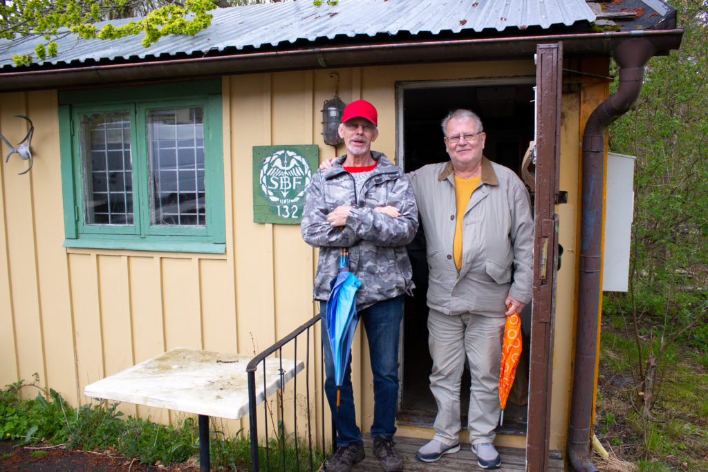 På bilden står Dennis och Lasse på trappen utanför den ljusgula stugan. Lasse håller en arm om Dennis axel. De både håller i varsitt paraply och de ler med stängd mun.