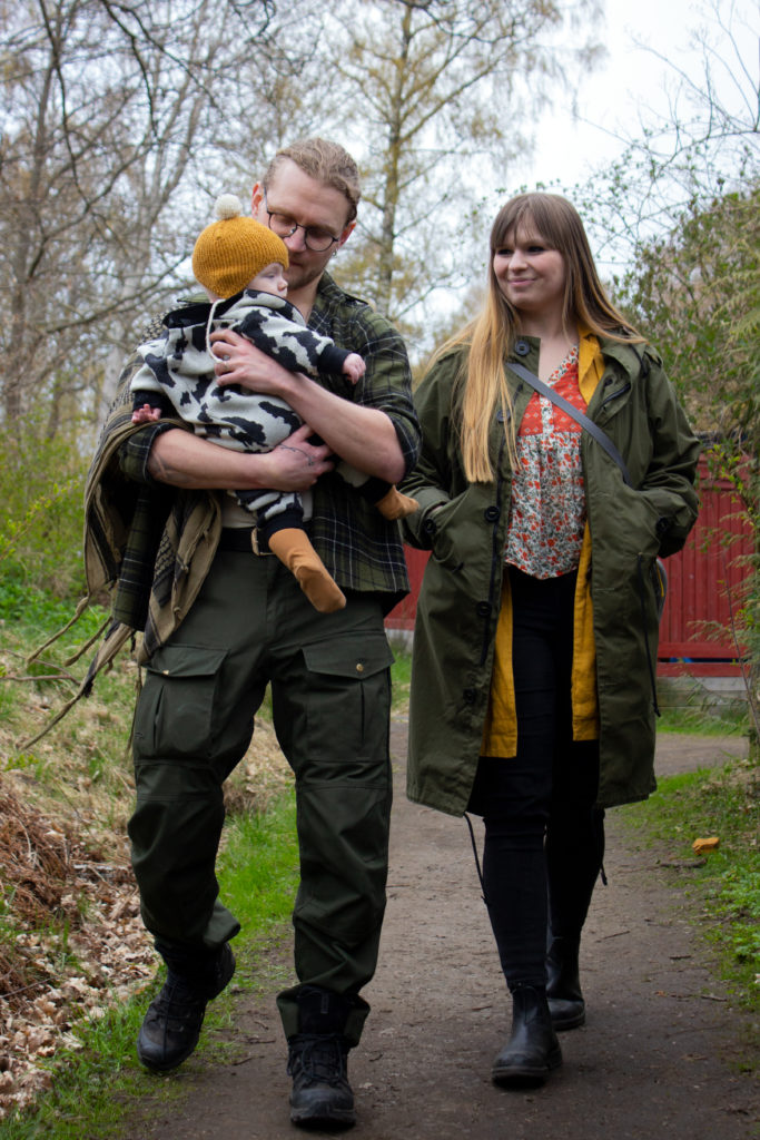 Fredrik och Tuva går på en stig i Sjöbergen. Bilden är en helkroppsbild där man ser de framifrån. Fredrik håller Aron i famnen. Aron har en gul, stickad mössa på sig.