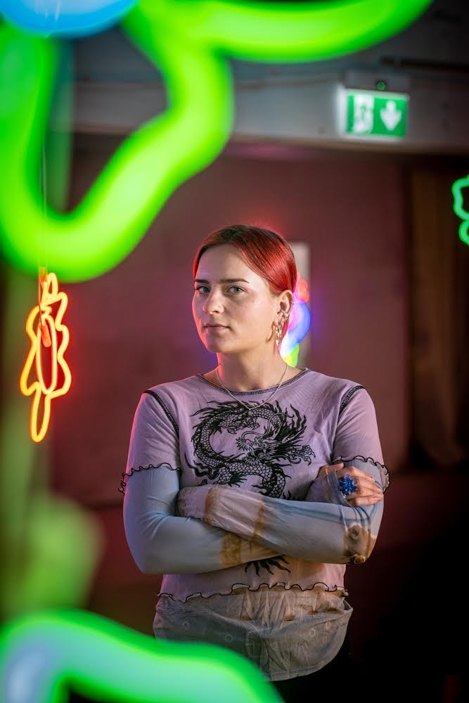 Porträtt av Josefin, från midjan och uppåt. Hon håller armarna i kors och sneglar in i kameran. Hon har på sig en lilla t-shirt med en svart drake på. Under t-shirten har hon en tröja med batikmönster. Hon har en stor blå ring på höger hand, örhängen i vänster öra och rött hår. Från vänster sida har hon ett rött sken i ansiktet från ett neonverk som hänger snett bakom henne. I förgrunden hänger fler av hennes neonverk, gröna och ur fokus.