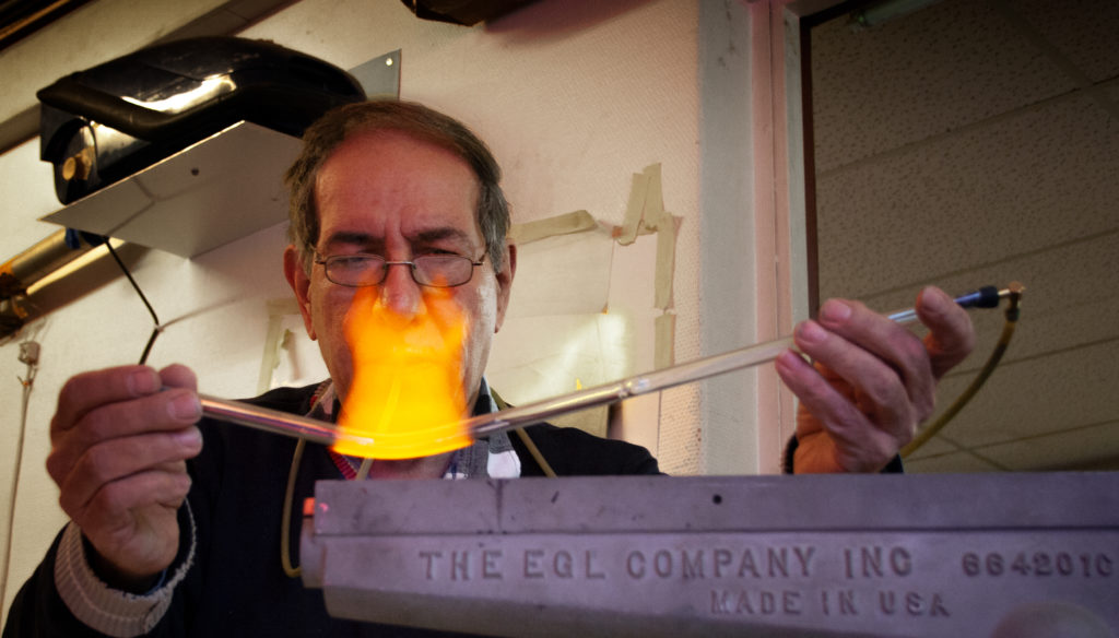 Ibrahim Hassan böjer ett glasrör i en låga från en av sina brännare. I förgrunden ser man brännaren med texten "THE EGL COMPANY INC 6642010 MADE IN USA". En stark gulorange låga flammar upp från ena kanten på brännaren. Genom lågan ser man Ibrahim som sitter bakom brännaren och tittar fokuserat på det smala glasröret som han håller i lågan med båda händerna. Han har fått till en lätt böj på glaset. I bakgrunden ser man ena väggen av hans arbetsrum. Uppe i vänstra hörnet har han en dammsugare uppe på en hylla.