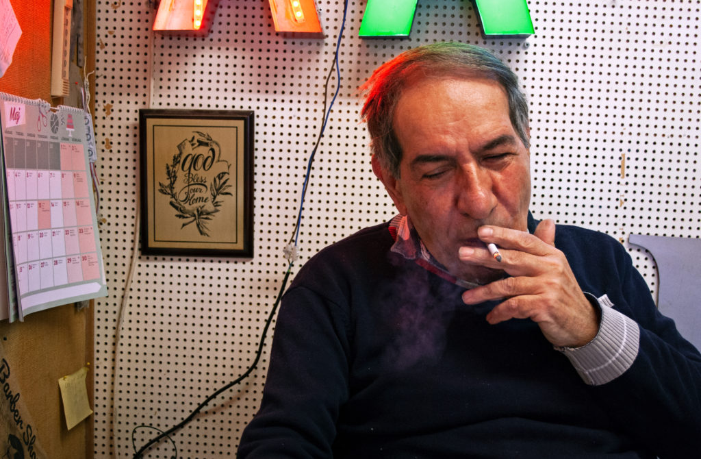 Halvbild på Ibrahim som tar ett bloss från sin cigarett. Det glöder från cigaretten och ett lätt rökmoln bildas strök under den. Ibrahim har svart tröja på sig. Han sluter ögonen och ser ut att njuta. Ovanför honom ser man skymten av två led-bokstäver som lyser i rött och grönt. De avger även ett rött och ett grönt sken på ibrahims hjässa. Till väsnter i bilden ser man en mässingsskylt med löv och snirkliga bokstäver: "God bless our home". En almacka hänger längst till vänster i bild.