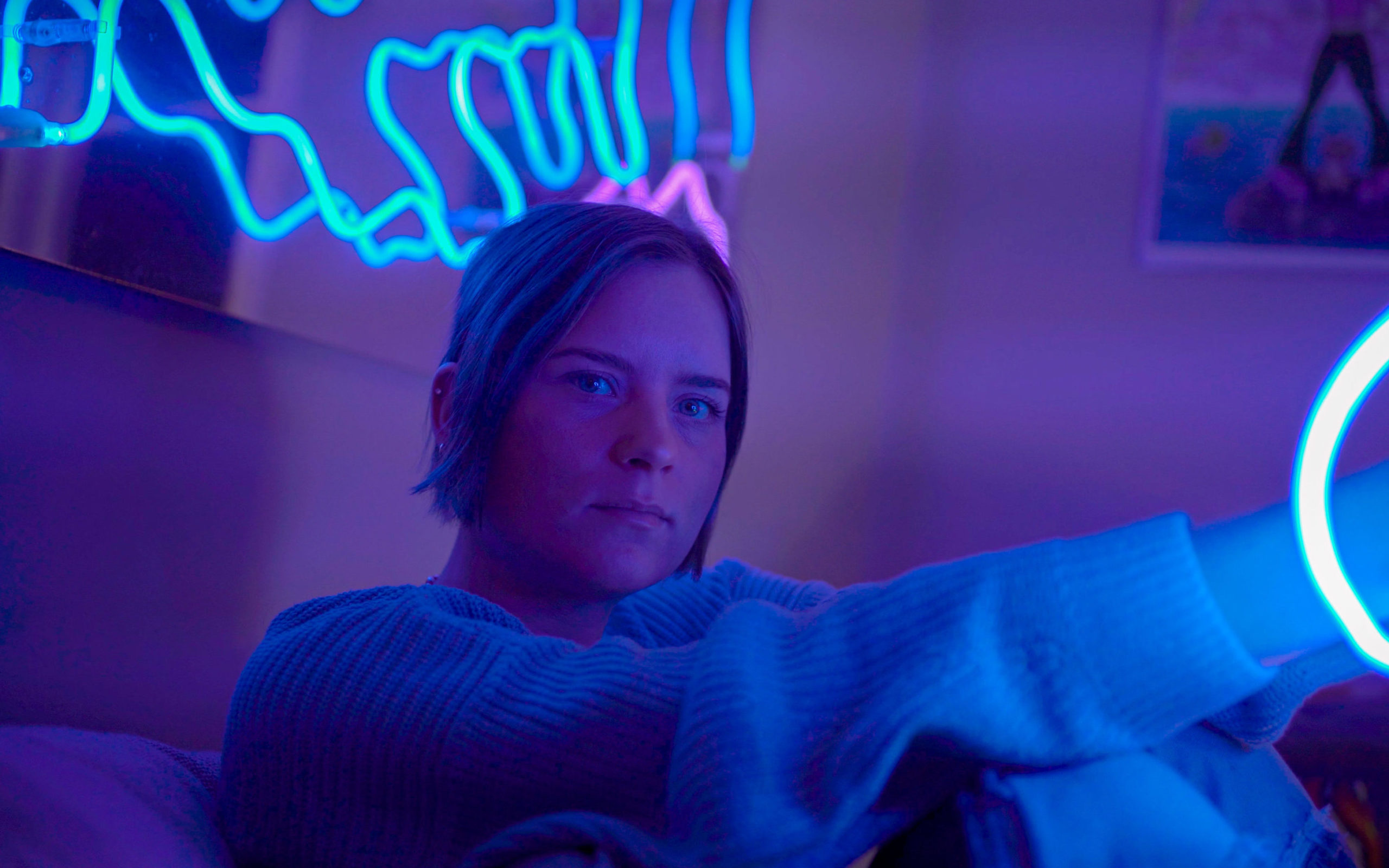 Stor bild på Josefin som sitter ner på någon slags soffa. Man ser bara hennes överkropp. Hon har armarna på sina knän. Hon tittar rakt in i kameran. I bakgrunden ser man en av hennes neoninstallationer; neonrör i blåa färger som har en snirklig form.