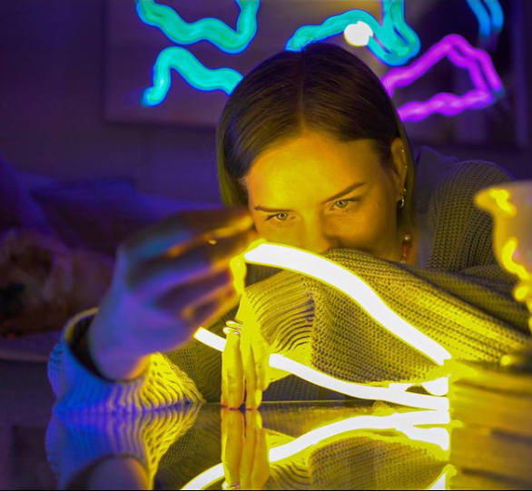 Rund bild på Josefin Eklund som har armarna uppe på en bordsskiva. Bordsskivan är blank och speglar Josefins armar och händer. I händerna håller hon ett neonrör som lyser starkt gult, skenet lyser upp hela hennes ansikte. Hennes blick är fäst på neonröret, medan hennes ena handled täcker hennes mun. I bakgrunden skymtar man ett mörkt rum som är upplyst av olika neonrör i färgerna blått och lila.