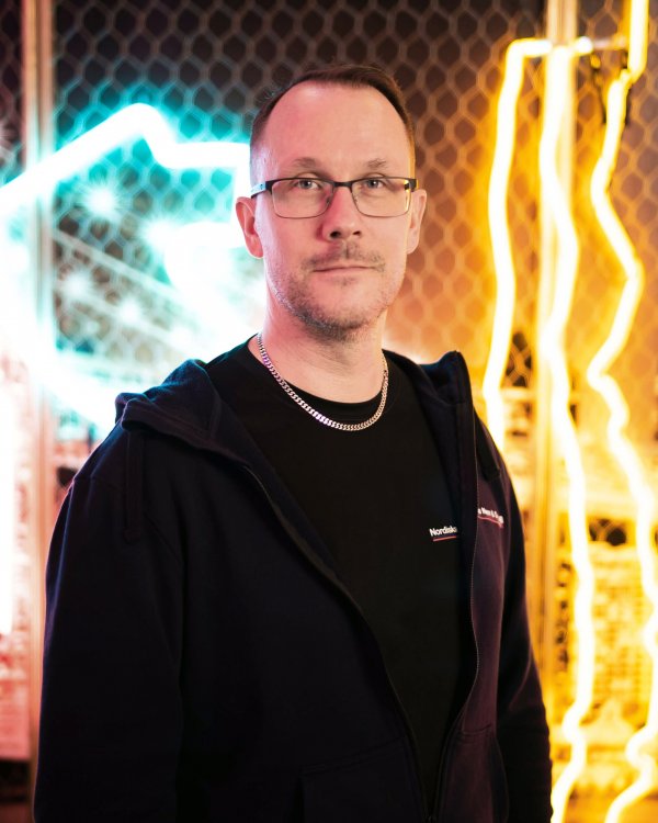 Porträttbild på Tommy Gustafsschiöld i halvfigur. Han har en svart tröja och ett guldhasband, samt ett par fyrkantiga glasögon. Han blickar rakt in i kameran. Bakom honom ser man olika neonljuskonstellationer; till vänster neonrör i turkos; och till höger neonrör i gult.